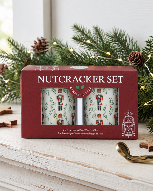 A red gift box labeled 'NUTCRACKER SET' sits on a white wooden mantel, surrounded by pine branches, pinecones, and a gold wick trimmer. The box has a clear window showing two patterned candles with nutcracker illustrations.