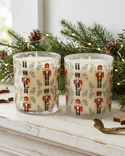 Two holiday-themed scented candles in clear glass holders with a cream wax base and a printed pattern of nutcrackers and evergreen branches, styled on a white wooden mantel with pine branches and pinecones.