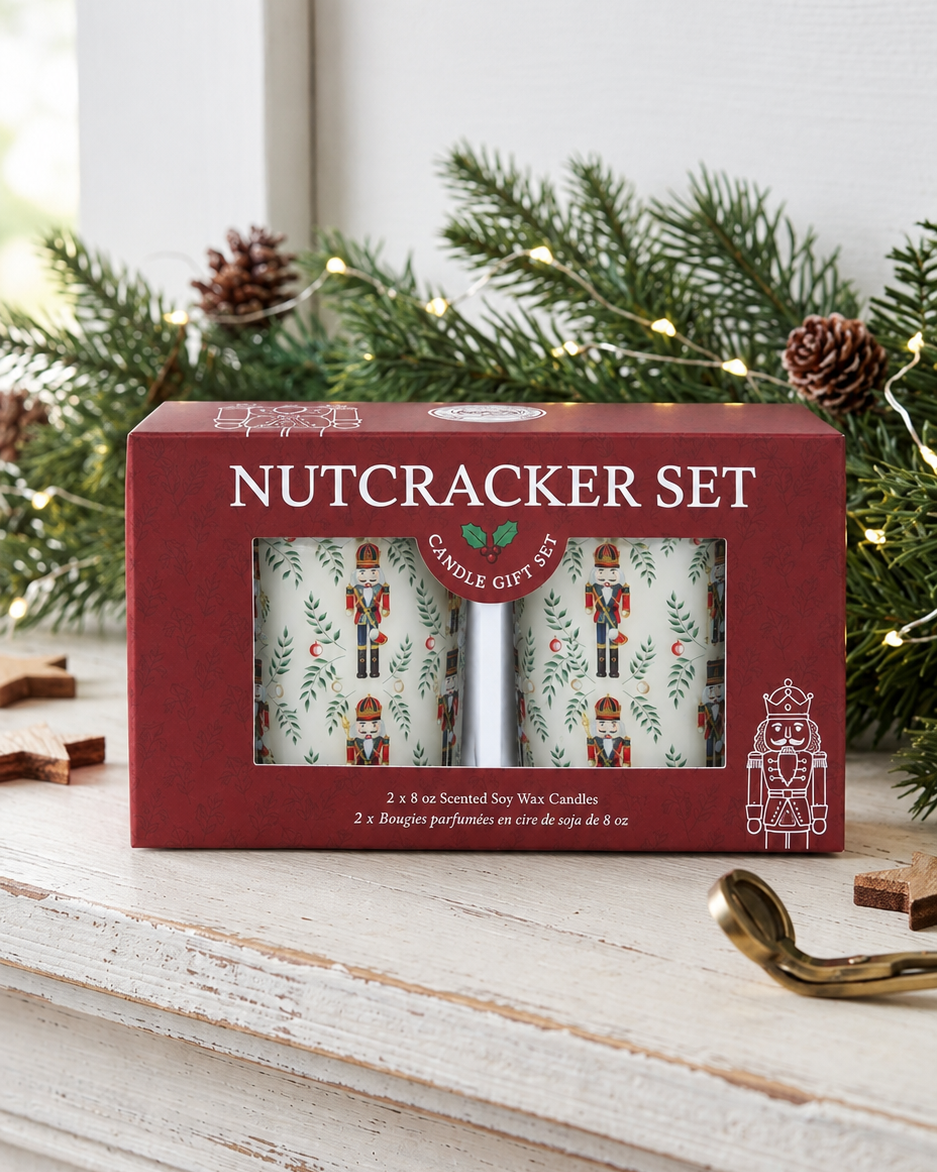 A red gift box labeled 'NUTCRACKER SET' sits on a white wooden mantel, surrounded by pine branches, pinecones, and a gold wick trimmer. The box has a clear window showing two patterned candles with nutcracker illustrations.