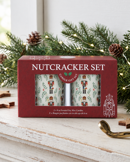 A red gift box labeled 'NUTCRACKER SET' sits on a white wooden mantel, surrounded by pine branches, pinecones, and a gold wick trimmer. The box has a clear window showing two patterned candles with nutcracker illustrations.