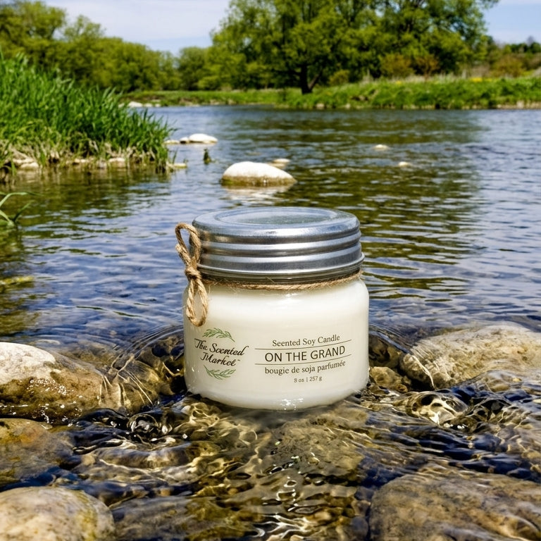 Jar of candle labeled 'On The Grand' in a river with greenery in the background